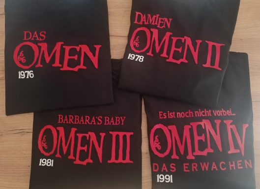 Vier schwarze T-Shirts mit roten Texten zu den Filmen "Das Omen" und Fortsetzungen.