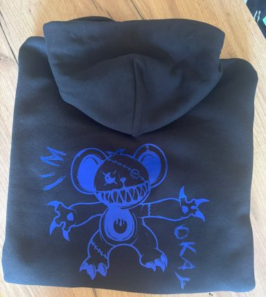 Schwarzes Hoodie mit blauer Zeichnung eines cartoonhaften Monsters und dem Text "OKAY".