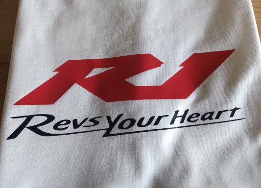 Logo mit den Buchstaben "R1" und dem Slogan "Revs Your Heart" in Rot und Schwarz.
